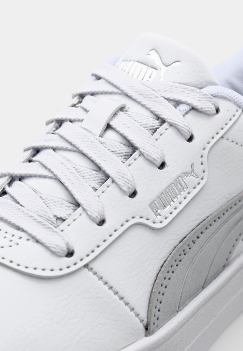 Sneaker en cuir blanc avec des accents gris, surface texturée, lacets plats et un logo argenté sur la languette. Vue en gros plan de la chaussure.