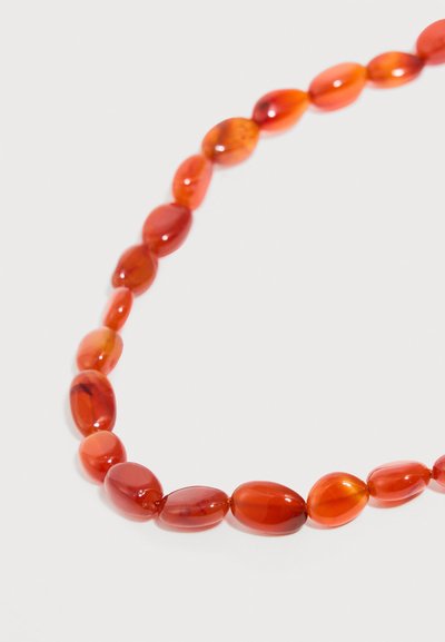 Chaîne de perles ovales irrégulières polies de couleur orange-rouge disposées en ligne courbe sur un fond blanc uni.