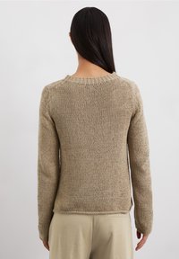 Femme aux longs cheveux noirs portant un pull en tricot beige et un pantalon beige, debout dos à l'appareil photo sur un fond blanc uni.