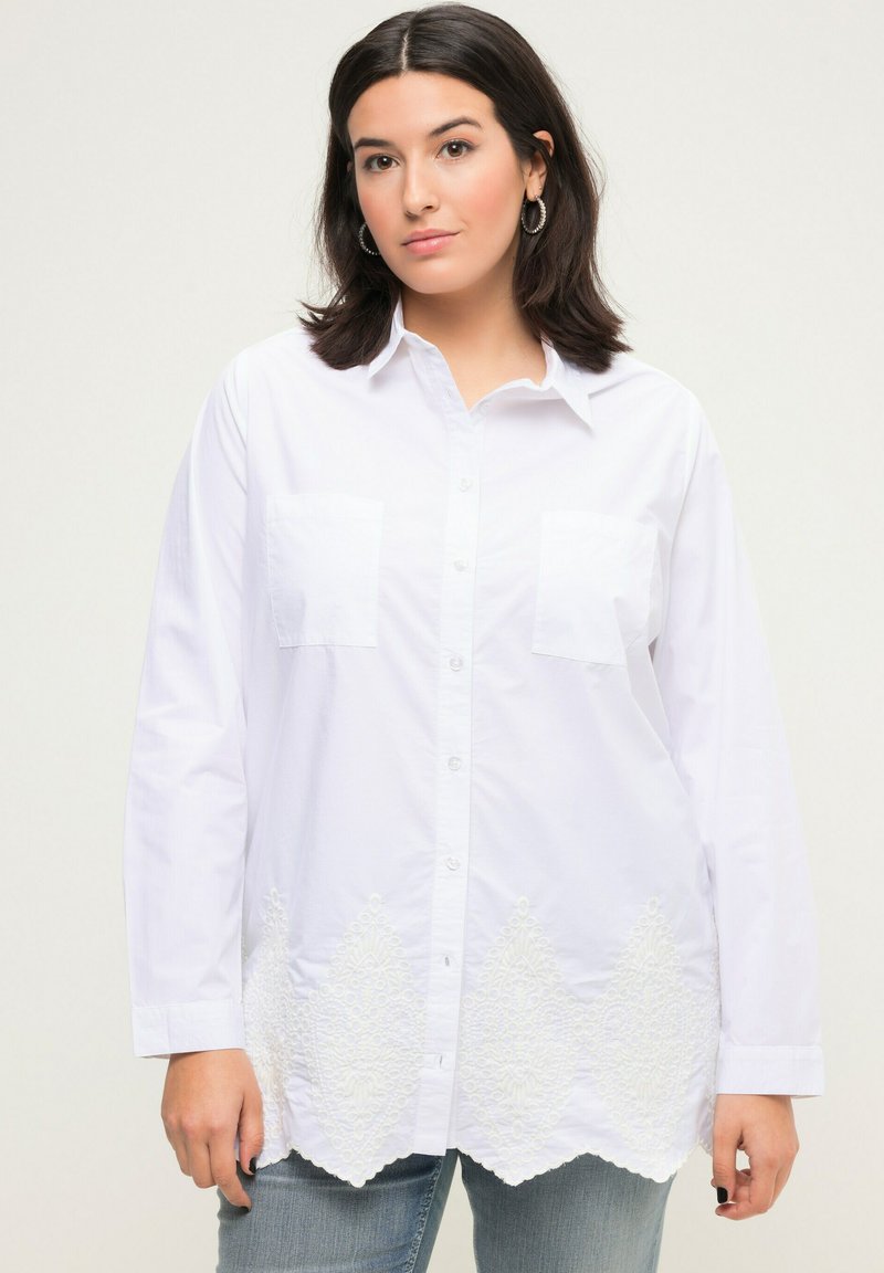 Camisa blanca de manga larga con cuello puntiagudo, bolsillos frontales y un intrincado bordado blanco en el dobladillo con un borde ondulado.