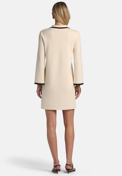 Žena v krémových šatách po kolená s čiernym lemovaním na golieri a manžetách, stojaca chrbtom, má na nohách čierne sandále na podpätku a zlaté náramky.