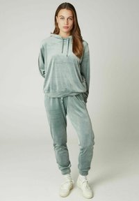 Samt-Jogginganzug in sanftem Grün, mit einer Kapuze zum Zuziehen, langen Ärmeln und schmal zulaufenden Hosen mit elastischen Bündchen. Weiße Sneakers vervollständigen das Outfit.