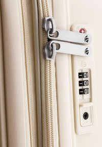 Cerniera color crema con doppi cursori metallici, accanto a un lucchetto a combinazione e a una serratura, su una valigia leggera e texture.