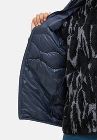 Reißverschlussjacke mit einem inneren Futter aus glattem, dunkelblauem Material und einer äußeren Schicht aus strukturiertem, schwarz-grauem Fleece mit Muster.