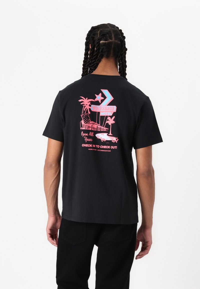 Personne avec des cheveux tressés portant un t-shirt noir avec un panneau de motel rose et bleu, des palmiers et un graphique de voiture vintage dans le dos.