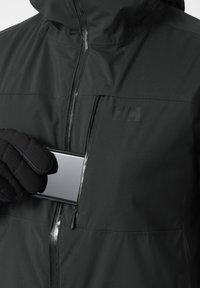 Zwarte geïsoleerde jas met een ritssluiting op de borstzak. Een hand in een zwarte handschoen houdt een smartphone dichtbij de zakopening.