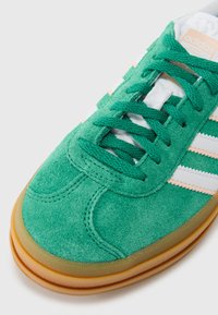 Πράσινο σουέντ αθλητικό παπούτσι με λευκές και ροδακινί λεπτομέρειες, στρογγυλεμένη μύτη, υφασμένα κορδόνια και σόλα από καουτσούκ gum.