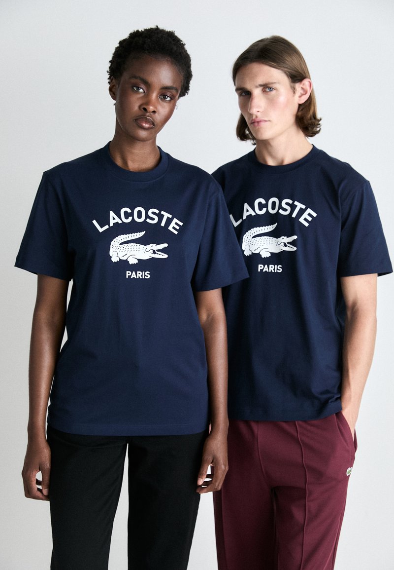 Tricouri din bumbac navy cu logo-ul alb "LACOSTE" și grafică cu crocodil, purtate cu pantaloni negri și bordo. Design clasic cu decolteu rotund.