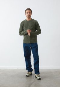 Khaki grøn strikket sweater med teksturerede mønstre og ribbede manchetter, parret med blå denim bukser og camouflaged sko.