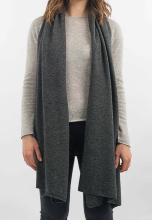 Femme portant un pull beige clair, une longue écharpe gris foncé et un pantalon noir, debout devant un fond clair uni.