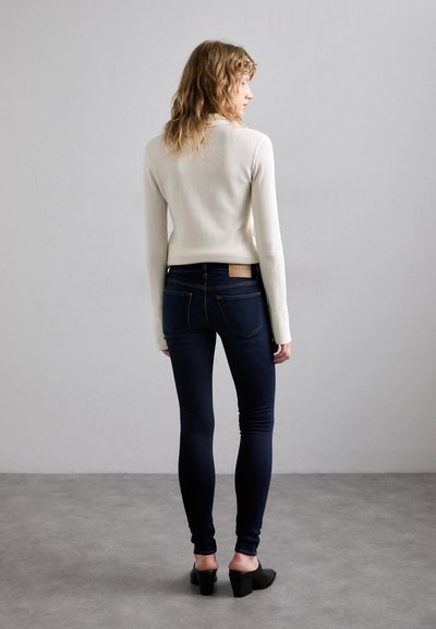 Crème ribbeltrui gecombineerd met donkerblauwe skinny jeans, voorzien van een leren label aan de achterkant, en zwarte hakken.