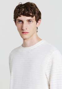 Maglione bianco con texture e scollatura rotonda, caratterizzato da un motivo sottile e una finitura morbida. Il modello che indossa il maglione è rivolto di fronte.