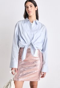Svetlomodrá košeľa s krátkym rukávom zviazaná v páse nad lesklou ružovou sukňou s flitrami. Model drží krémovú tašku.