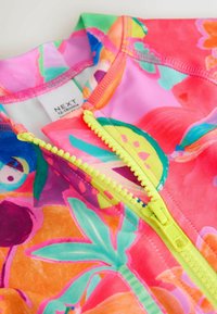 Rash guard színes virágmintával rózsaszín, zöld és kék árnyalatokban. Jellemzője egy sárga cipzár és puha, sima anyag. Címke látható belül.