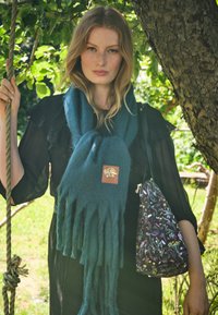 Tyrkysová šatka s třásněmi a koženou nášivkou, nosená s čiernymi šatami; drží kvetinovou kabelku pred prírodným pozadím.