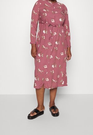 Šaty s dlouhým rukávem v barvě prachové růže s květinovým potiskem, zapínáním na knoflíky vpředu, páskem v pase a kulatým spodním okrajem. Nošeny s černými pantoflemi.