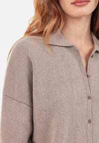 Cardigan tricoté marron clair avec col, doté d'une patte de boutonnage et d'une texture douce. Comprend des variations subtiles de couleur.