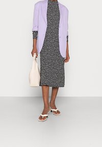 Persona vestida con un vestido midi de flores en blanco y negro, blazer lila, sandalias de tacón blancas, sosteniendo un bolso blanco frente a un fondo liso.