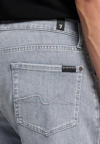 Jeans en denim gris clair vus de dos avec une poche arrière visible, dotés de surpiqûres blanches et d'une petite étiquette noire attachée.