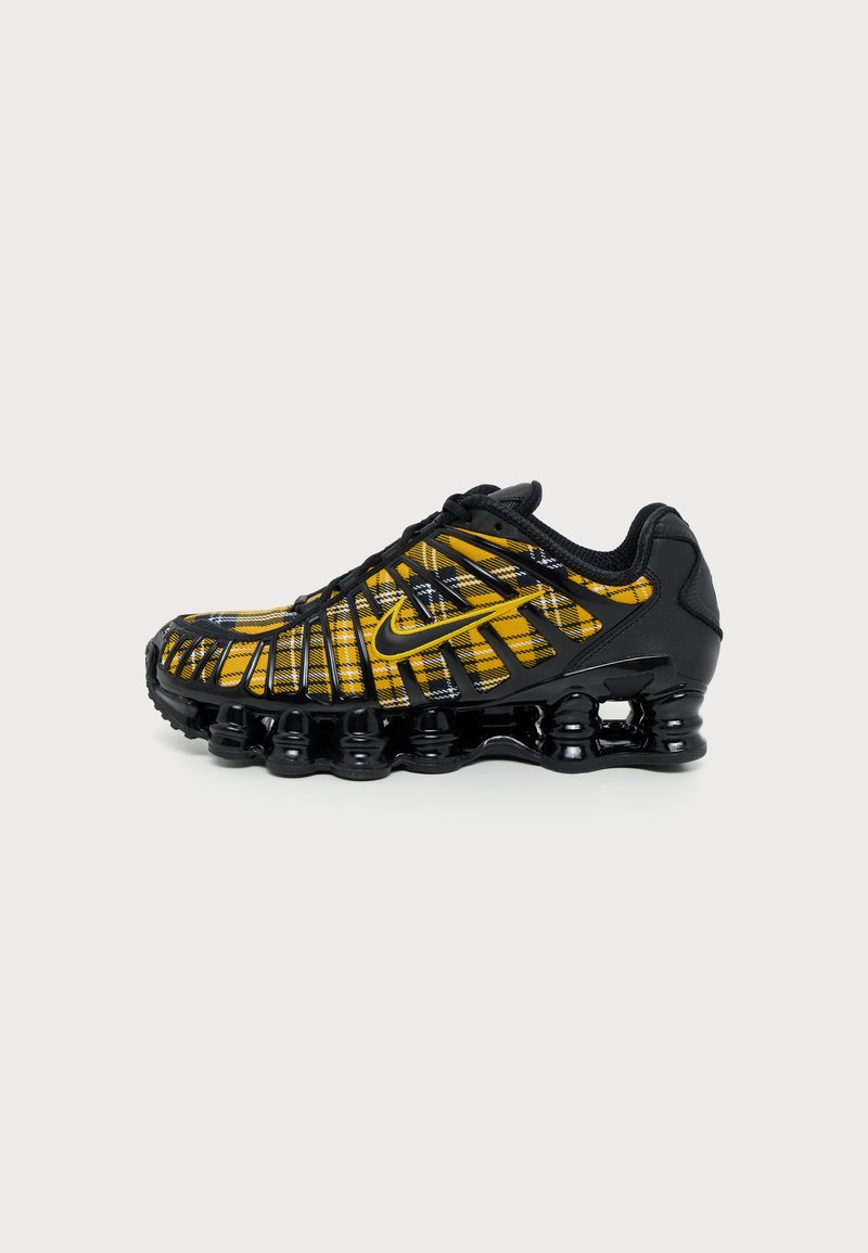Μαύρο και κίτρινο αθλητικό παπούτσι Nike με καρό μοτίβο, ορατή αερόσολα και μαύρα κορδόνια, εμφανίζεται από το πλάι σε λευκό φόντο.