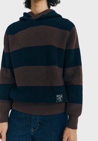 Personne portant un hoodie tricoté à rayures larges bleu marine et marron avec une petite étiquette noire, associé à un jean bleu foncé.