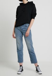 Felpa nera con cappuccio e tasca a marsupio, abbinata a jeans a gamba dritta blu chiaro. Calzature: sneakers alte nere e bianche.