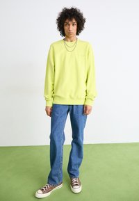 Neon grøn sweatshirt med rund halsudskæring, lange ærmer, ribbede manchetter og kant, kombineret med blå straight-leg jeans og brune sneakers.