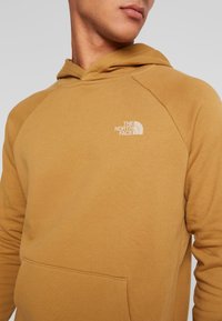 Sudadera con capucha marrón hecha de tela suave, que presenta un bolsillo tipo canguro y un pequeño logo blanco en la zona del pecho.