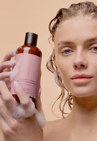 Brun flaska med ett svart lock, märkt "Ceramide Complex Bond Repair Shampoo." Håret är blött med skum. Ljus bakgrund.