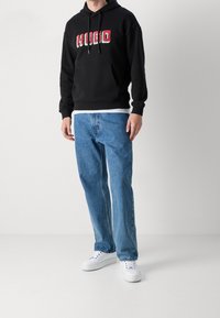 Felpa nera con logo "HUGO" colorato, abbinata a jeans in denim azzurro e sneakers bianche. Presenta una tasca frontale e cappuccio con coulisse.