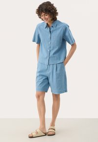 Hellblaues, gestreiftes Kurzarmhemd mit einer Brusttasche, kombiniert mit passenden Shorts. Das Material wirkt atmungsaktiv und eignet sich für warmes Wetter.