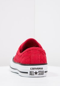 Zapatilla de lona roja con textura tejidos, diseño bajo, suela de goma blanca, acentos en negro y marca "Converse All Star" en el talón.