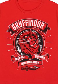 Rød bomulds T-shirt med et grafisk motiv af et løvehoved, omgivet af teksten "Gryffindor" og bånd med teksten "Mod", "Dristighed" og "Determination".