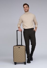 Hombre sonriente vestido con un suéter de cuello alto beige y pantalones oscuros, sostiene el asa de una maleta dura dorada con cuatro ruedas.