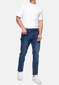 Hombre vistiendo una camiseta blanca de manga corta simple, jeans ajustados azul y zapatillas blancas, posando frente a un fondo claro y liso.