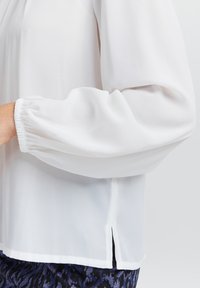 Hvid, let bluse med lange, samlede ærmer og en sideslids i kanten. Stoffet har en glat, gennemsigtig tekstur.