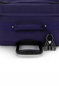 Lila Reisetasche mit einem ausziehbaren schwarzen Griff, Textur aus Stoff, einem Stoffgurt und einem schwarz-weißen Gepäckanhänger in Form einer Figur.