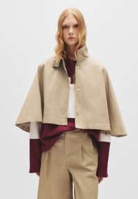 Giacca cropped beige con cappotto e alto colletto, con bottoni a pressione e una texture liscia, indossata sopra un maglione bordeaux e bianco.