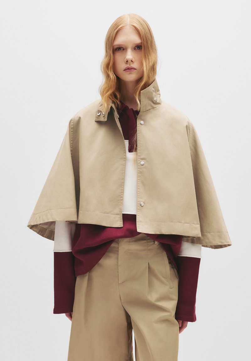 Giacca cropped beige con cappotto e alto colletto, con bottoni a pressione e una texture liscia, indossata sopra un maglione bordeaux e bianco.