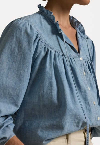 Világoskék chambray blúz fodros gallérral, gombos záródással, valamint ráncolt anyaggal a vállaknál és az ujjaknál, lazán illeszkedő fazonnal.