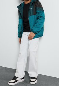 Farverig vindtæt jakke i teal og sort, sort t-shirt, hvide vide jeans og sort-hvide sneakers med snørebånd.