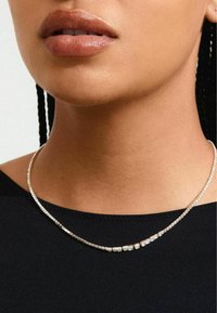 Hopeaketju hopeasta, jossa useita pieniä, kimaltelevia yksityiskohtia, käytetään paljaalla kaulalla mustan paidan päällä. Silkkisen tasainen ja heijastava pinta.