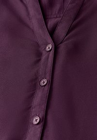 Blouse violette avec un décolleté en V et six boutons ronds. Texture lisse avec un éclat subtil, dotée d'une seule poche poitrine.
