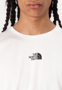 Bílý bavlněný tričko s černým logem značky The North Face v levém horním rohu. Krátké rukávy a kulatý výstřih.