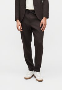 Personne portant un pantalon marron foncé sur mesure avec un blazer assorti et des baskets blanches à semelles en caoutchouc, debout devant un fond uni.