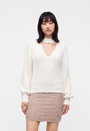 Femme aux cheveux noirs raides portant un pull blanc à col en V avec une découpe et une mini-jupe rose clair scintillante.