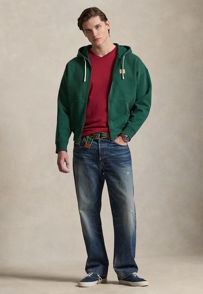 Νέος άνδρας που φοράει πράσινο φούτερ με φερμουάρ, κόκκινη μπλούζα με V και στενή λαιμόκοψη, μπλε τζιν, ριγέ ζώνη και μπλε αθλητικά παπούτσια, στέκεται μπροστά σε μπεζ φόντο.