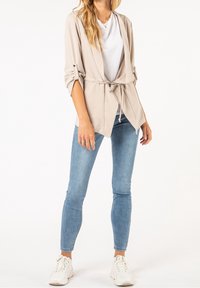 Beiger Blazer mit Gürtel und hochgekrempelten Ärmeln, getragen über einem weißen T-Shirt. Kombiniert mit hellblauen Skinny-Jeans und weißen Sneakers.