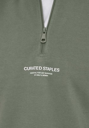 Felpa verde oliva con zip e collo alto, caratterizzata da testo stampato bianco "CURATED STAPLES" e un robusto dettaglio della cerniera.