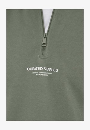 Sweat-shirt zippé vert olive avec un col haut, portant le texte imprimé en blanc "CURATED STAPLES" et un détail de fermeture éclair robuste.